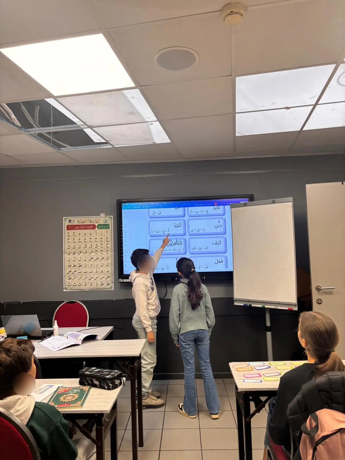 Deux enfants au tableau numérique pendant un cours d'arabe, devant l'alphabet affiché au mur